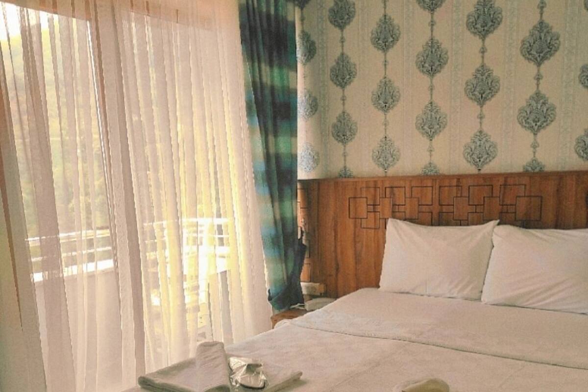 Una habitación de hotel con una cama bien hecha, una mesita de noche con lámpara y una ventana con cortinas translúcidas.