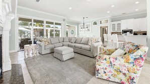 Un salon spacieux avec un canapé d’angle, un fauteuil à motifs floraux et un coin repas doté de meubles blancs, avec une cuisine en arrière-plan.