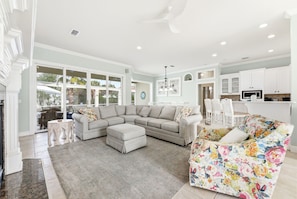 A spacious living room with a sectional sofa, a floral armchair, and a dining area with white cabinets and a kitchen in the background.