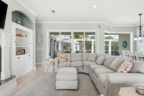 A spacious living room with a sectional sofa, a small ottoman, and a dining area with white chairs.