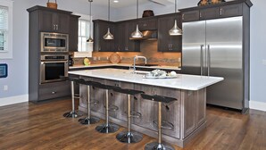 A modern kitchen with dark cabinetry, stainless steel appliances, a central island with bar stools, pendant lighting, and a large refrigerator. - Dream Weaver - 1232968 (Santa Rosa Beach)