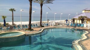 Une piscine aux eaux cristallines, dont les bords sont ondulés, entourée de chaises longues et de palmiers, avec une plage et l’océan en arrière-plan.