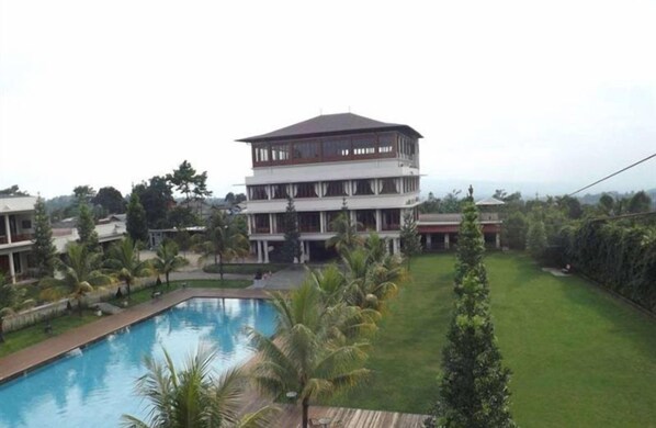 A multi-story hotel with a swimming pool, surrounded by well-maintained lawns and palm trees. - Jimmers Mountain Resort (Megamendung)