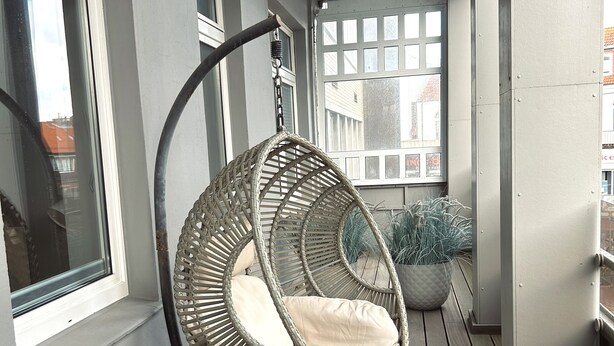 Una silla colgante de mimbre con cojines sobre una terraza de madera, una cesta de metal y una planta en maceta.
