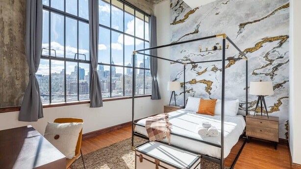 A modern bedroom with a glass four-poster bed, a city view, and a marble-patterned wall. - Sosuite | 2BR Penthouse w W/D, Gym, On-Site Bar (Philadelphia)
