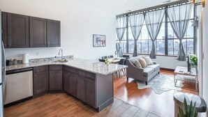 A modern kitchen with dark cabinets, a marble island, and a dining area with a gray sofa and city view. - Sosuite | 2BR Penthouse w W/D, Gym, On-Site Bar (Philadelphia)