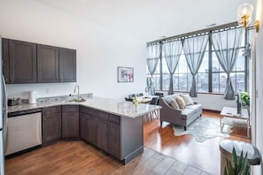 A modern kitchen with dark cabinets, a marble countertop, and a city view through large windows. - Sosuite | 2BR Penthouse w W/D, Gym, On-Site Bar (Philadelphia)