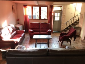 A living room with a sofa, armchairs, and a coffee table. There is a staircase to the right and a door leading outside.