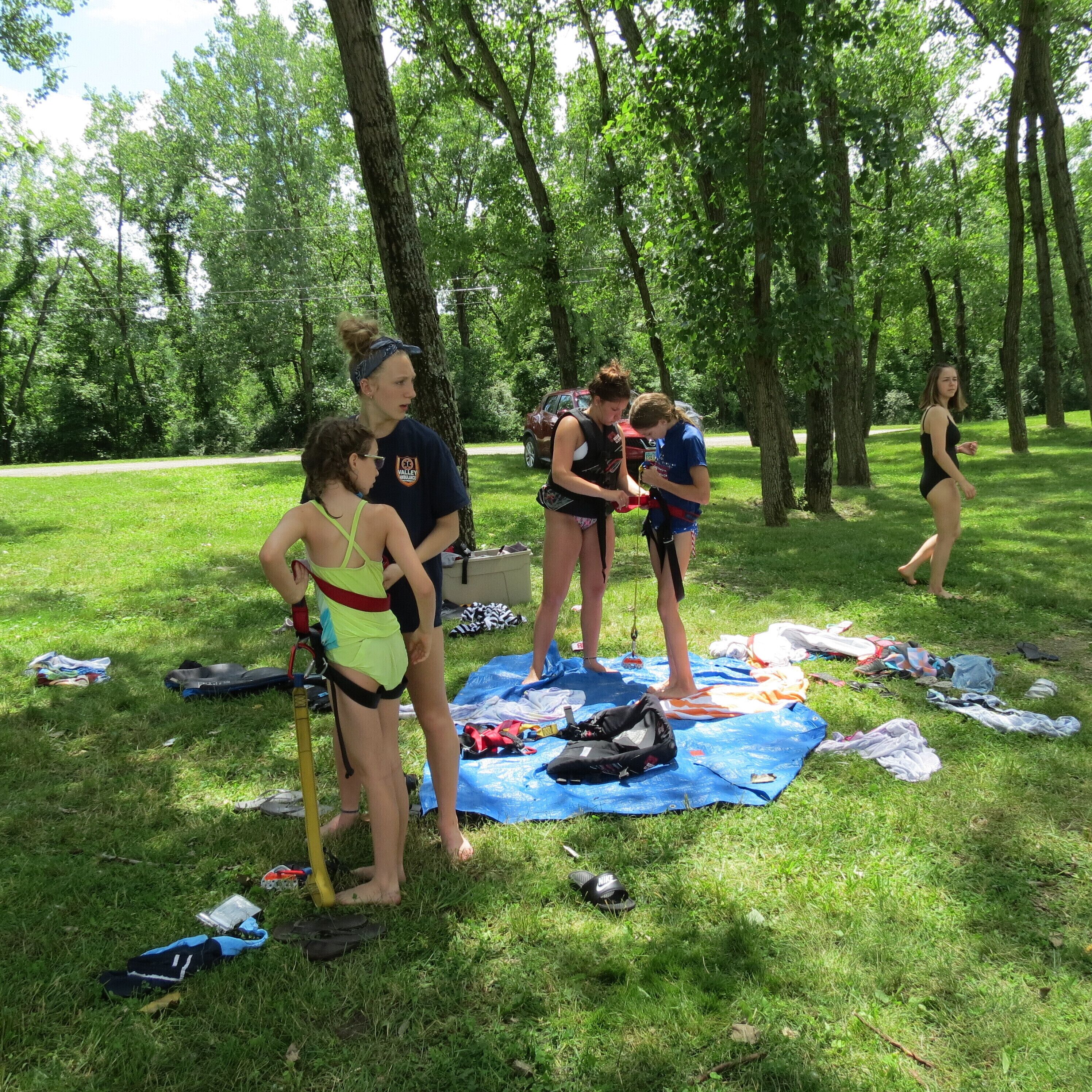 Un groupe de personnes se trouve dans un parc, avec des arbres, de l’herbe et un tapis bleu posé sur le sol.
