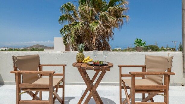 A wooden table with two chairs, a pineapple, and a plate with food and drinks on a rooftop terrace with a palm tree and a view of the sea. - Casa Verde di Naxos (Naxos)