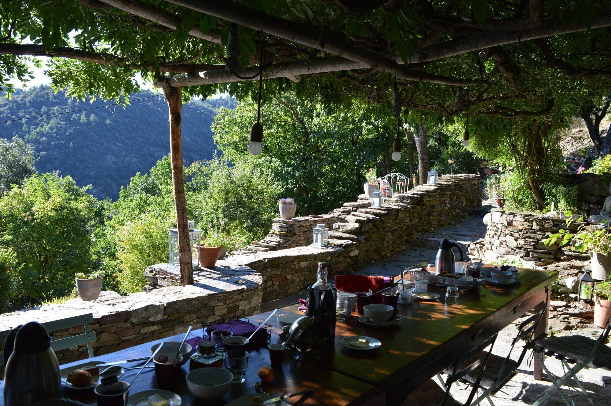 An outdoor dining area with stone walls and potted plants, overlooking a mountainous landscape.