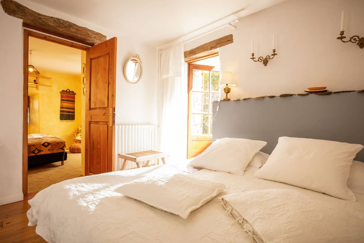 A bedroom with a large bed, wooden beams, a small table, and a window with curtains.