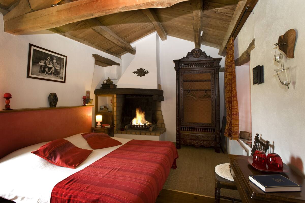 A cozy bedroom with a fireplace, a bed with red bedding, and a wooden ceiling.
