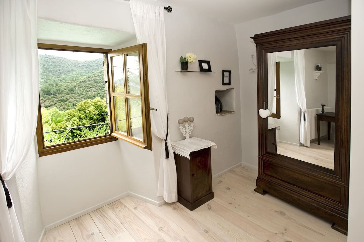 A bedroom with a large window, a wooden dresser, a mirror, and a shelf with decorative items.