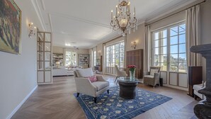 Un salon spacieux agrémenté d’un lustre, de grandes fenêtres et d’une cheminée.