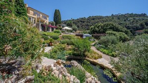 Un jardin luxuriant avec un petit étang, un bâtiment doté d’un balcon et une vue sur une colline parsemée d’arbres.
