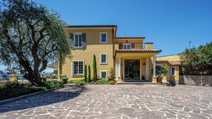 Un bâtiment jaune à deux étages, avec une entrée pavée de pierres, un grand arbre à gauche et un ciel bleu dégagé.