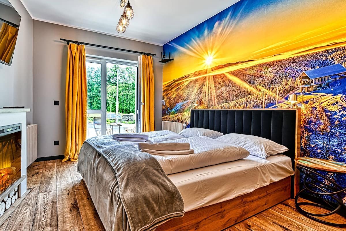Ein Schlafzimmer mit einem großen Bett, einem Kamin, einem Fenster mit Vorhängen und einem Wandgemälde von einem Sonnenuntergang.