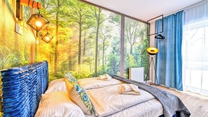 Une chambre avec une grande fenêtre, un lit avec une literie bleue et beige, et une fresque représentant une forêt sur le mur.