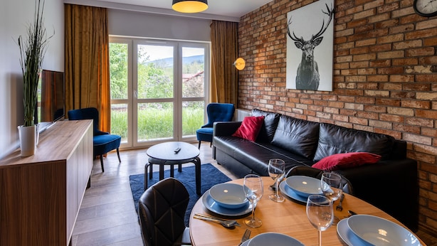 Un salon moderne avec un mur en briques, un canapé en cuir noir, une table à manger pour quatre personnes et une grande fenêtre donnant sur un paysage verdoyant.
