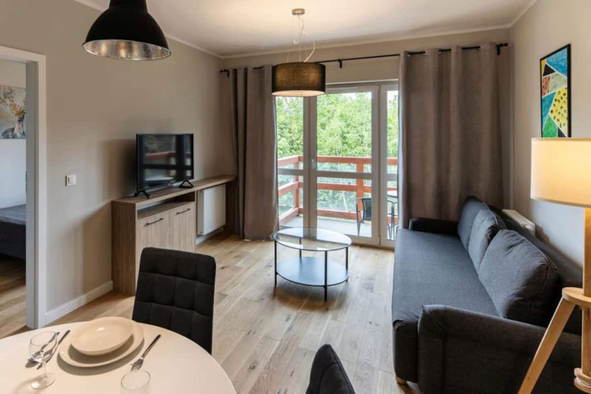Ein modernes Wohnzimmer mit einer Couch, einem Esstisch, einem Fernseher und einem Balkon mit Blick auf Grünflächen.