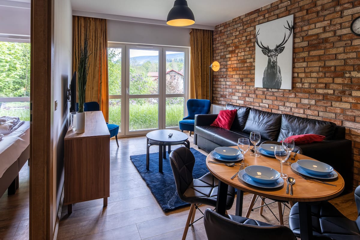 Ein modernes Hotelzimmer mit Backsteinwand, großem Fenster, Essbereich mit rundem Tisch und einer Couch mit Kissen.
