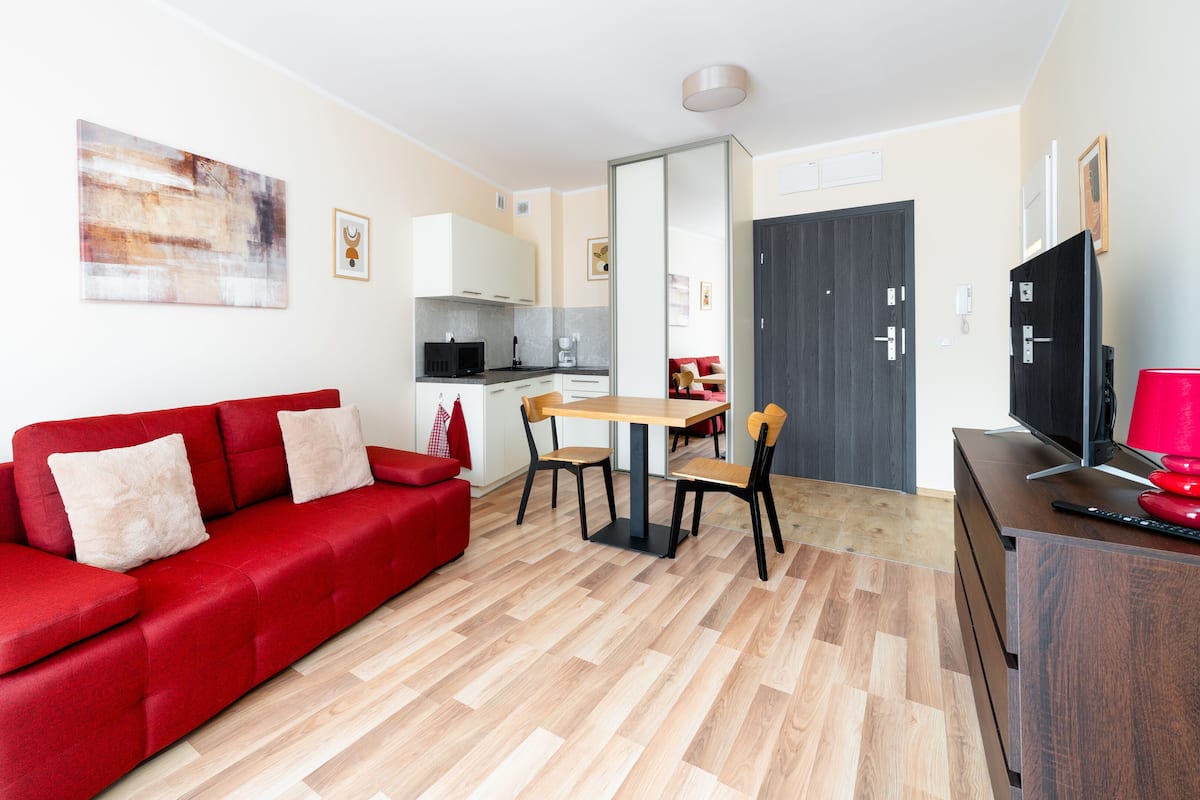 Ein kompakter Wohnbereich mit rotem Sofa, Esstisch mit Stühlen, Küchenzeile und Fernseher auf einem Holzkasten.