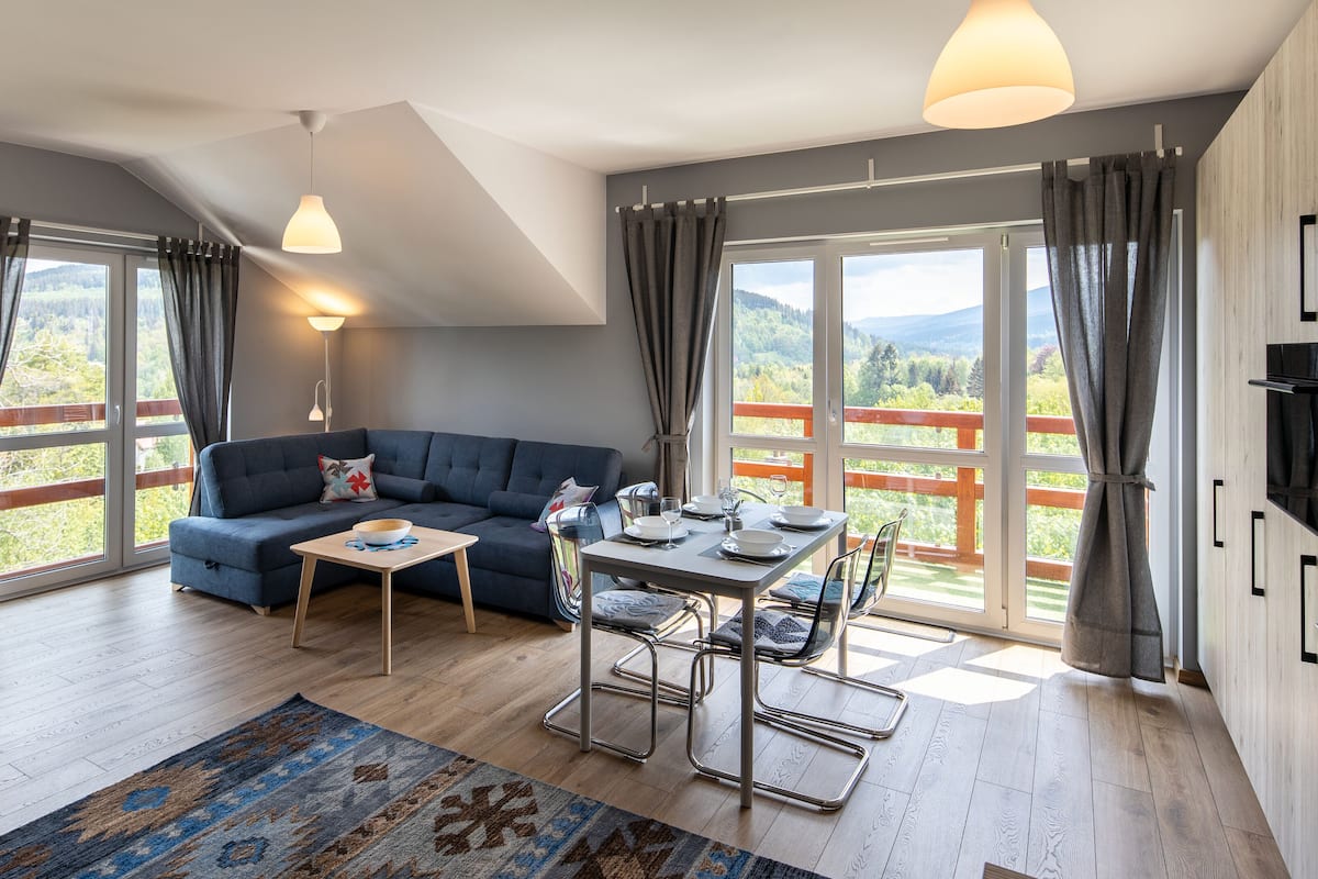 Ein modernes Wohnzimmer mit einer Couch, einem Esstisch und Stühlen, großen Fenstern mit Vorhängen und Blick ins Freie.