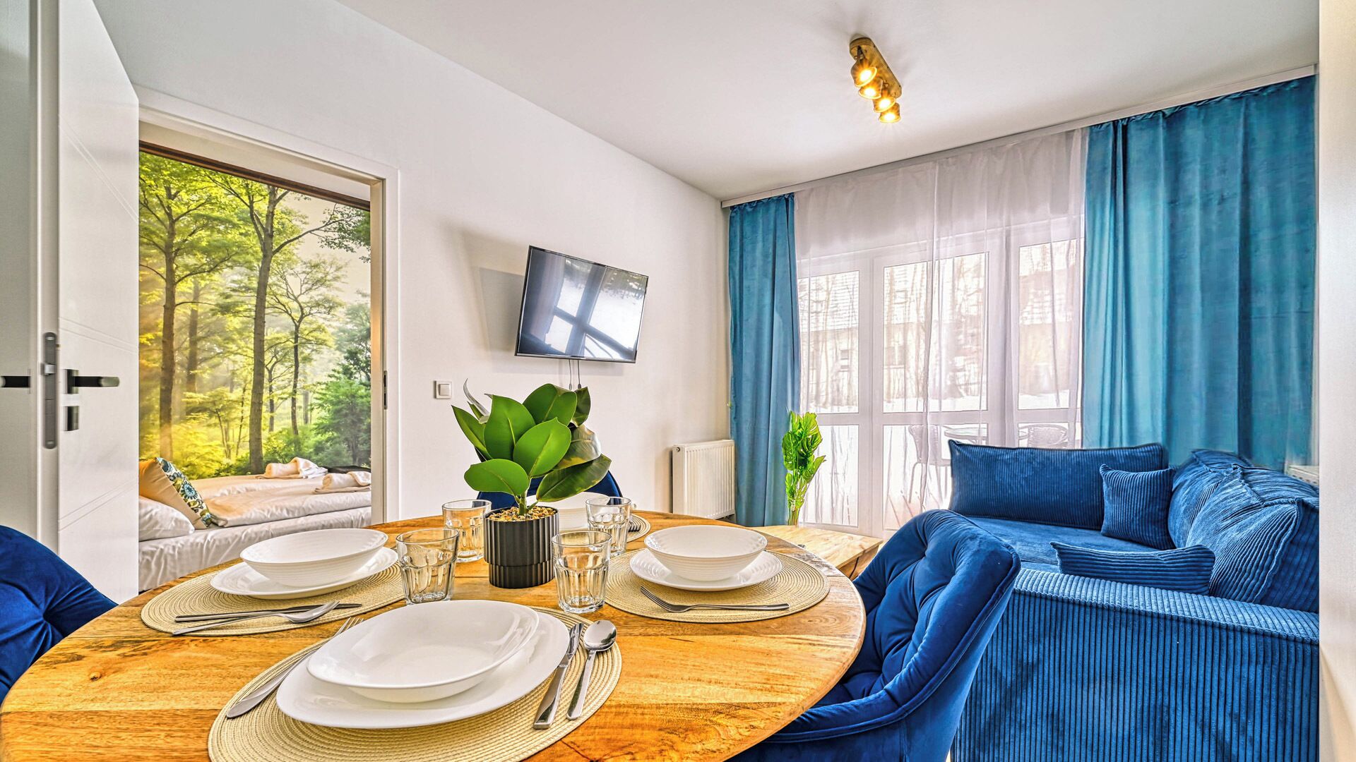 A modern living room with a dining table set for four, a sofa, a TV mounted on the wall, and a view of a forest through the window.