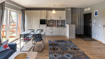 Una cocina moderna con armarios blancos, un comedor con mesa dispuesta para cuatro personas, y una sala de estar con sofá azul y alfombra estampada.