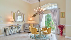 A dining area with a round table, chairs, and a chandelier. There is a sofa and a window with curtains.