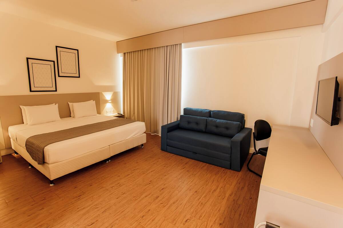 A hotel room with a bed, a sofa, a TV, and a window with curtains.