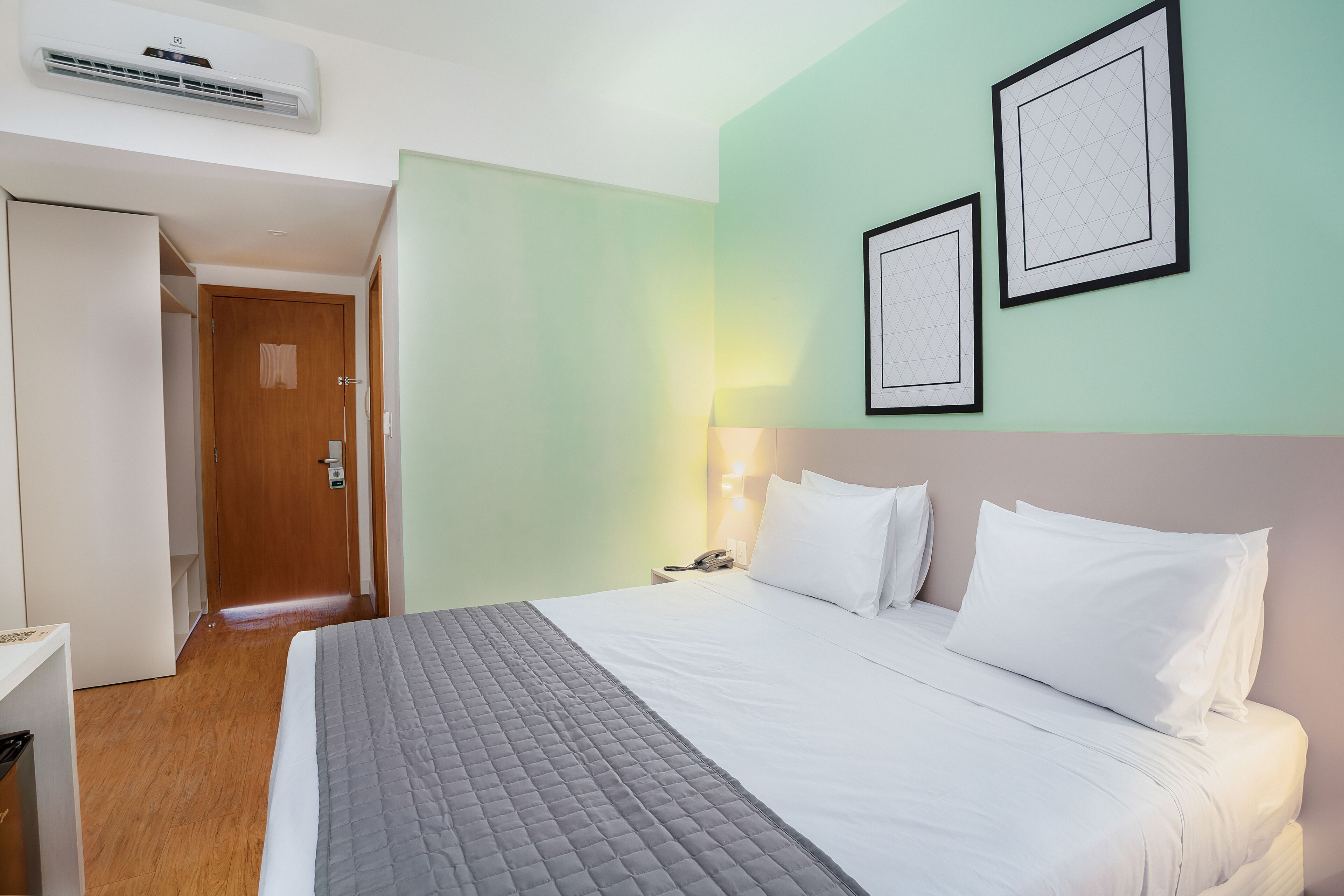 A hotel room with a bed, a bedside table, a lamp, and a wall-mounted air conditioner.