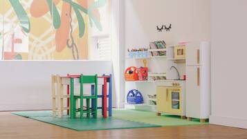 A child's playroom with a wooden floor, colorful chairs, a small kitchen set, and a window with a view of trees.