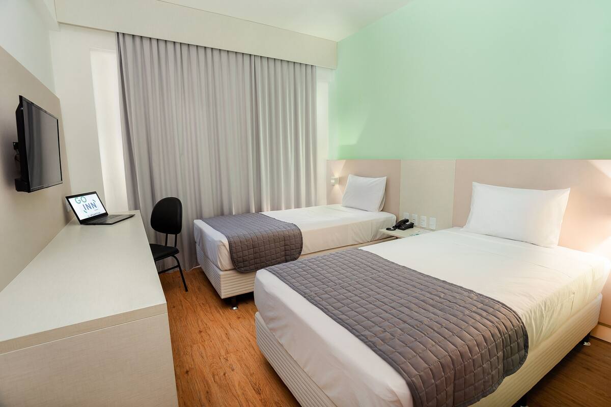 A hotel room with two beds, a desk with a laptop, a chair, and a TV on the wall.