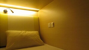 Una habitación pequeña, con poca luz, que tiene una cama individual, una lámpara de noche y un interruptor de luz en la pared.
