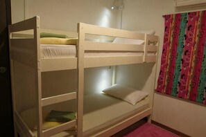 Una cama litera con un almohadón y una manta en la cama superior, un pequeño estante abajo y una ventana con cortina.