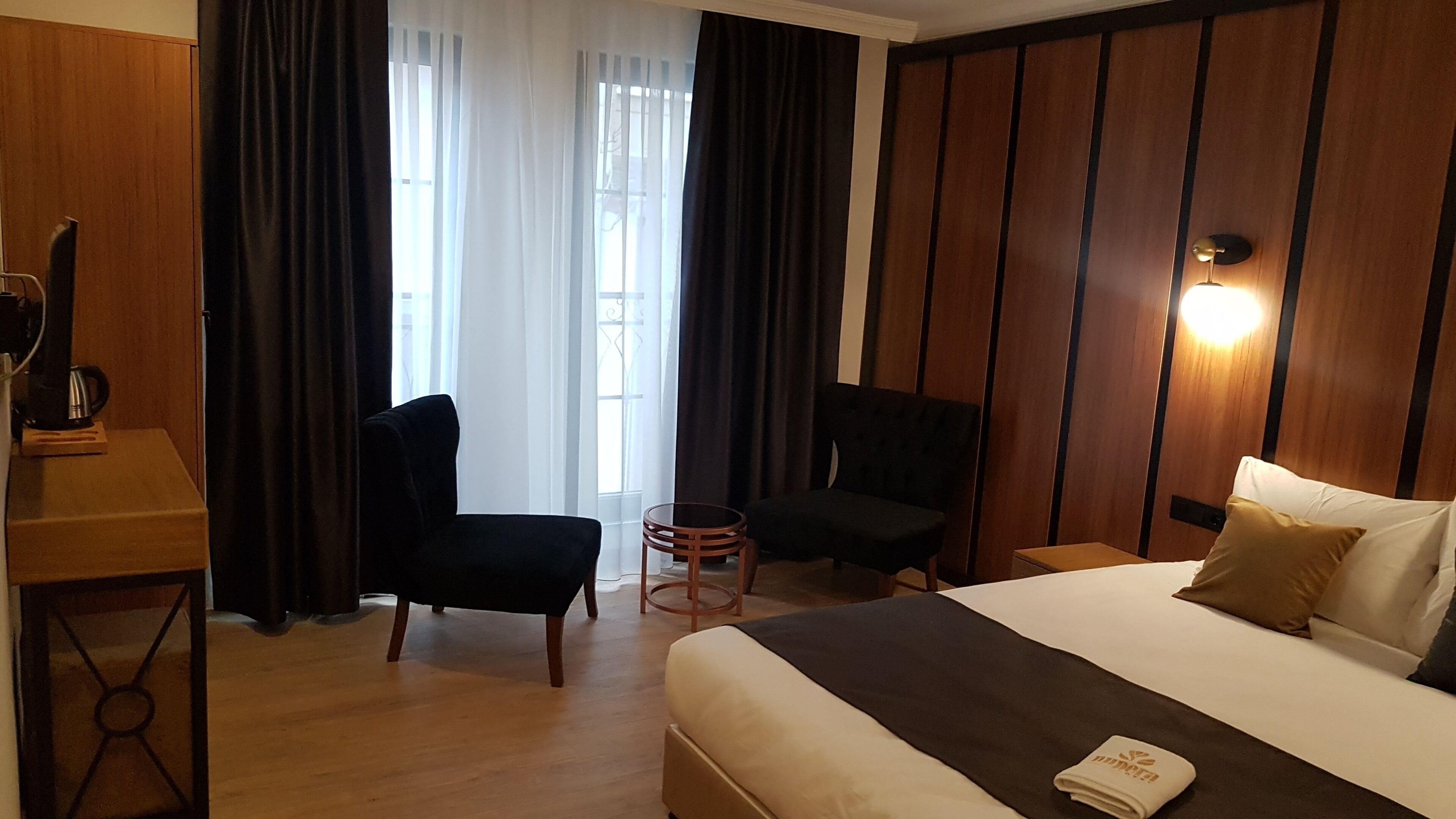 A hotel room with a bed, two chairs, a small table, a coffee machine, and a window with curtains.