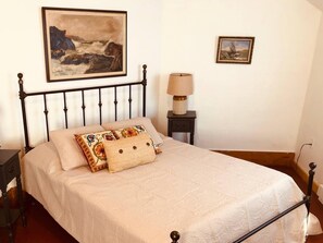 A bed with a white bedspread, a decorative pillow, and a headboard. There is a nightstand with a lamp and a small drawer. Two framed paintings are on the wall.