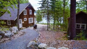 Un chalet rustique, accessible par un chemin de gravier, entouré d’arbres et de rochers.