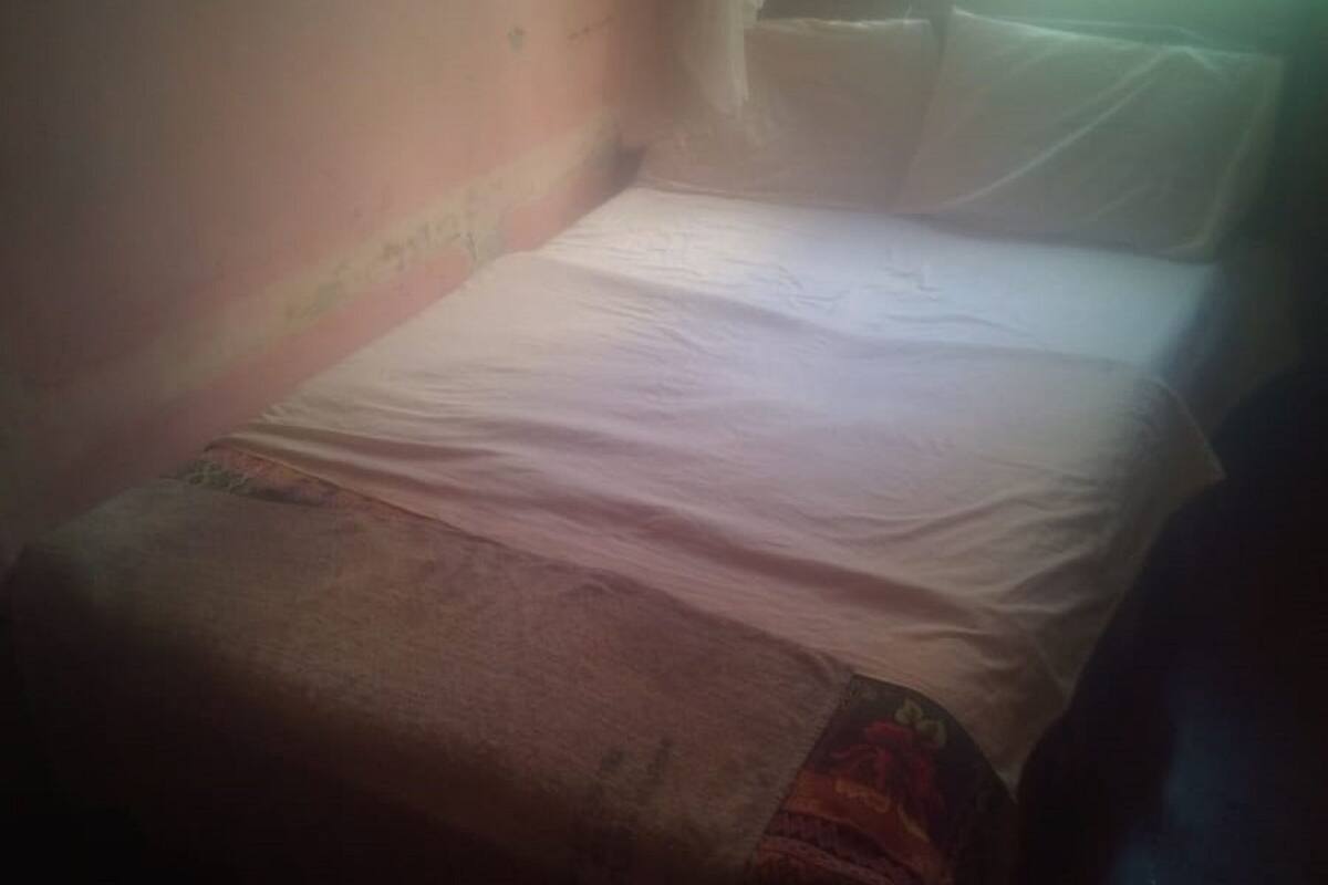 A dimly lit room with a single bed, a window with curtains, and a wall with a visible crack.