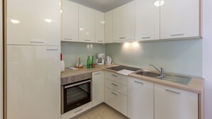 A compact kitchen with white cabinets, a built-in oven, a sink, and a countertop with a kettle and bottles.