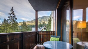 A balcony with a view of a forested mountain, a wooden railing, and a glass door leading to a room with a table and chairs.