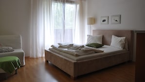 A bedroom with a bed, a chair, a bedside table, and a window with sheer curtains.