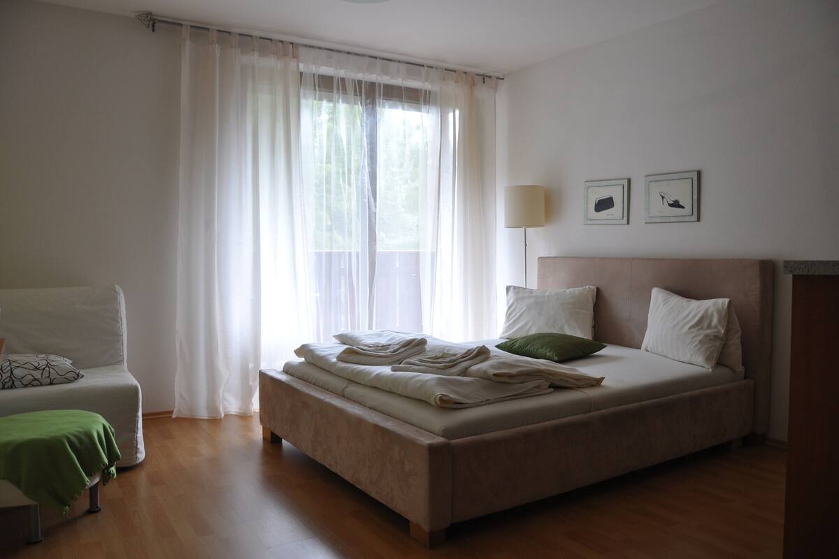 A bedroom with a bed, a chair, a bedside table, and a window with sheer curtains.