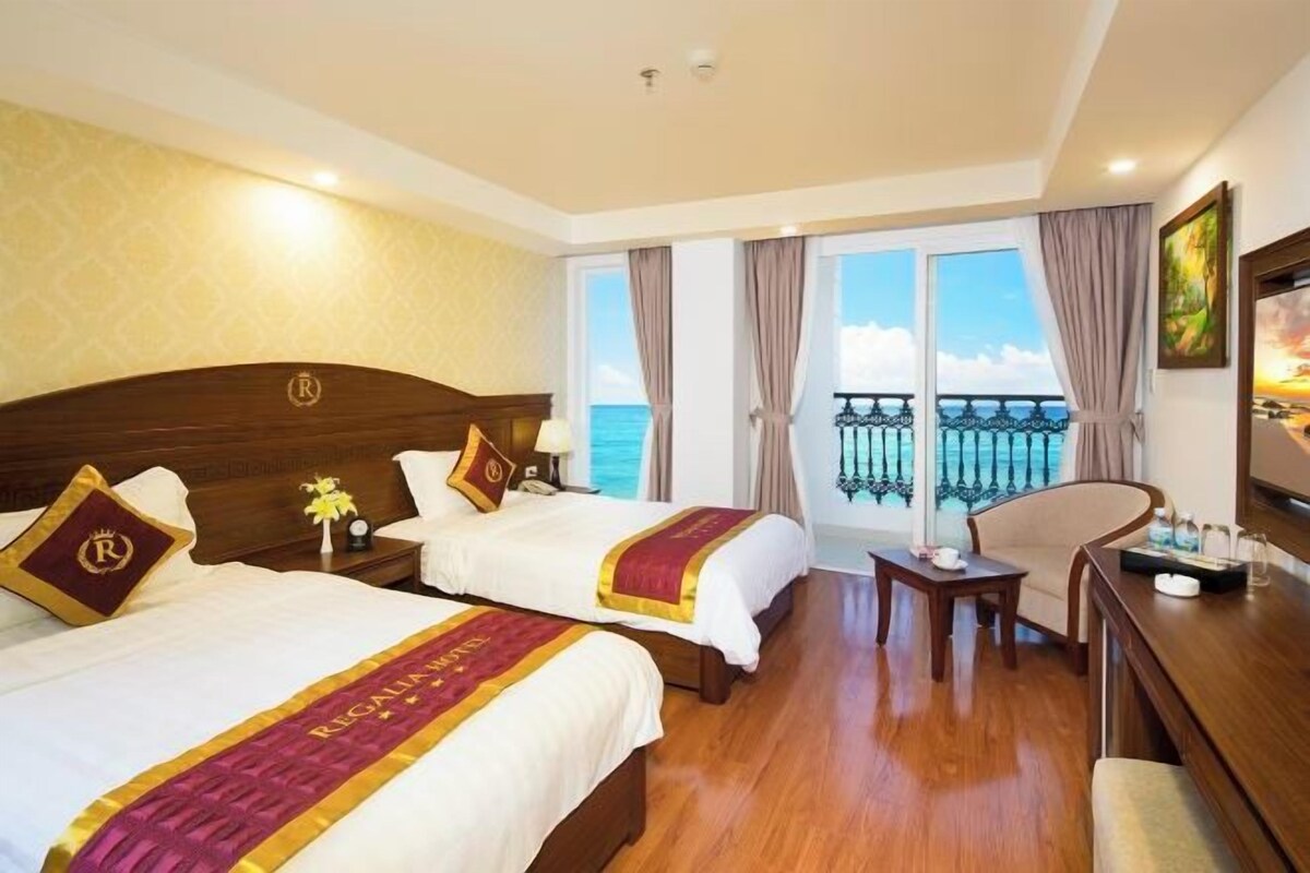 Habitación de hotel con dos camas, un balcón con vista al mar y un escritorio con una silla.