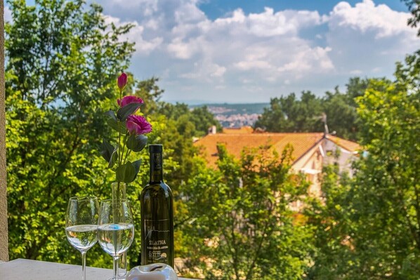 Een tafel met een fles wijn, twee glazen en een bloemenboeket, met uitzicht op een pittoresk landschap met huizen en bomen.