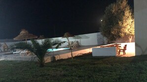 Área externa à noite com piscina, espreguiçadeiras e mesa de jantar.
