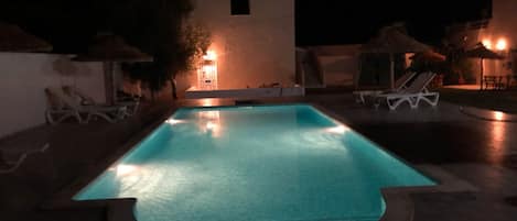Una piscina nocturna con superficie de agua cristalina, rodeada de sillas de descanso y sombrillas.