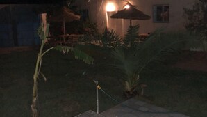 Espaço ao ar livre à noite com um pátio ladrilhado, um guarda-sol de palha e um edifício com uma janela iluminada.